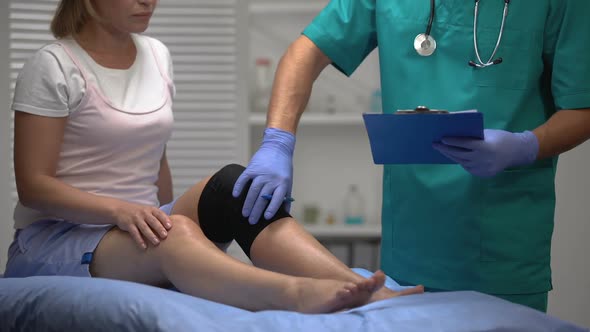 Orthopedist Checking Female Patient Leg With Knee Brace and Filling Document