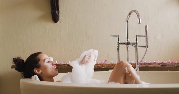 Happy biracial woman lying in bathtube, blowing foam and pampering herself with rose petals