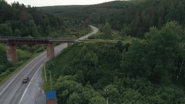 An Abandoned Rusty Railroad Bridge Above the Curving Road with a Driving Truck