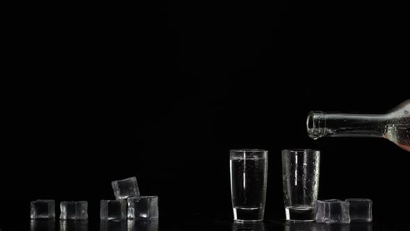 Hand Pours Vodka Tequila or Sake From Bottle Into Shot Glasses on Black Background with Ice Cubes