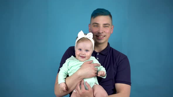 A Young Father with Blue Hair Holds a Baby in His Arms. Alternative Man with a Child Look at the