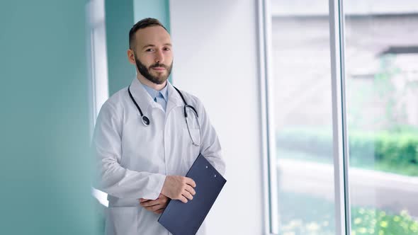 Handsome Confident Doctor in White Coat Looking at Window and Holding Clipboard in His Hands