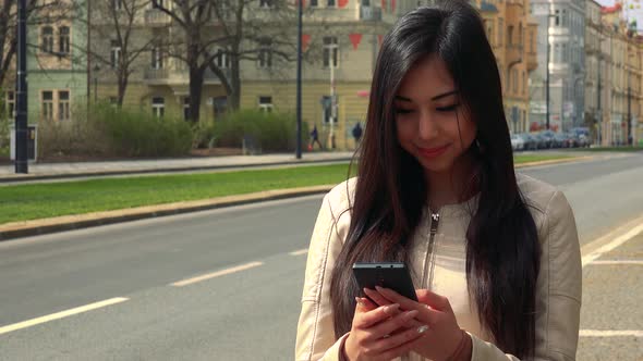 A Young Asian Woman Works on a Smartphone with a Smile in a Street in an Urban Area