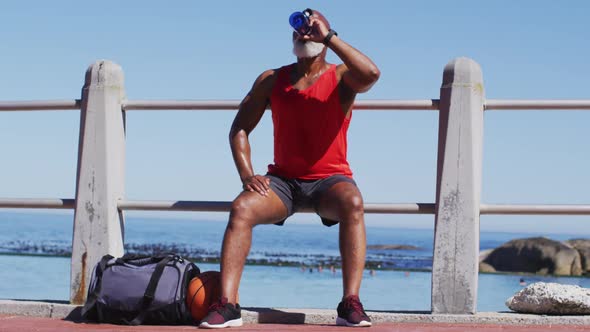 Senior african american man drinking water while sitting on the railing near the beach