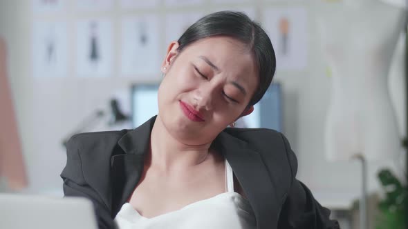 Asian Female Designer In Business Suit Working On A Laptop And Stretching While Designing Clothes
