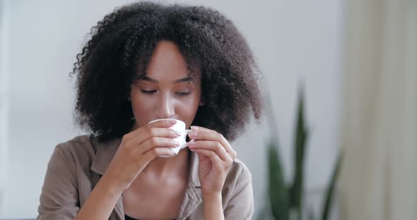 Smiling Happy African American Woman Sitting on Sofa in Casual Clothes, Picking Up Hot Cup of Coffee