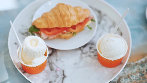 The Girl Takes the Ice Cream Off the Plate Next to the Croissant with the Meat