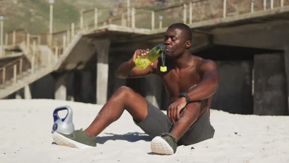 Tired african american man sitting on beach, drinking from bottle, taking break in exercise outdoors