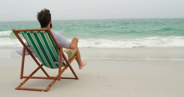 Rear view of man relaxing with hands behind head on sun lounger at beach 4k