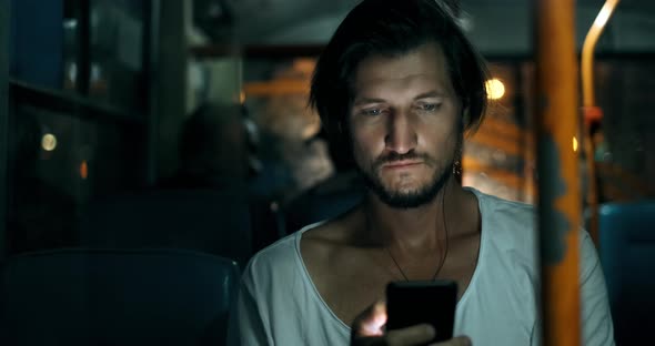 Young Man Holds Smartphone While Traveling By Bus at Night. Bearded Male in Headphones with Phone