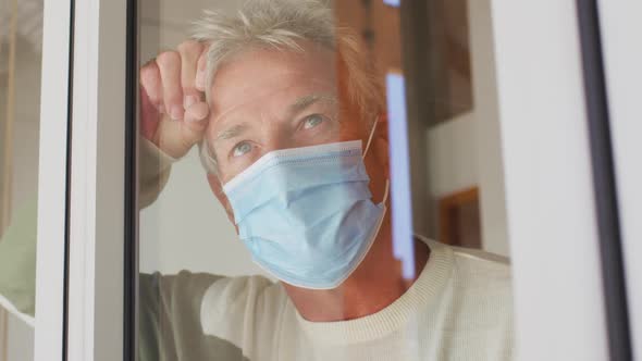 Senior caucasian man wearing face mask looking out of window at home