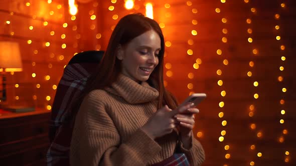 Happy Woman in Winter Sweater Typing Online Message on Mobile Phone, Sitting at Rocking Armchair.