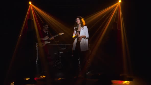 A Stylish Girl Vocalist Sings on Stage in a Vintage Microphone. In the Background Musicians