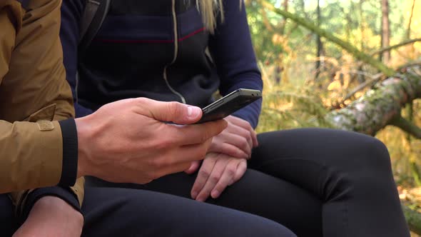 A Hiking Couple Sits on a Broken Tree in a Forest and Works on a Smartphone - Closeup