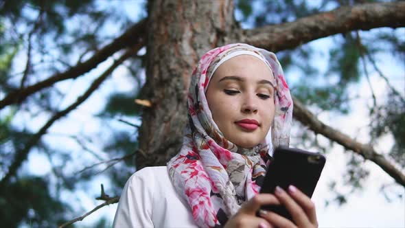 A Muslim in Hijab Communicates with Friends on a Mobile Phone in a Summer Park