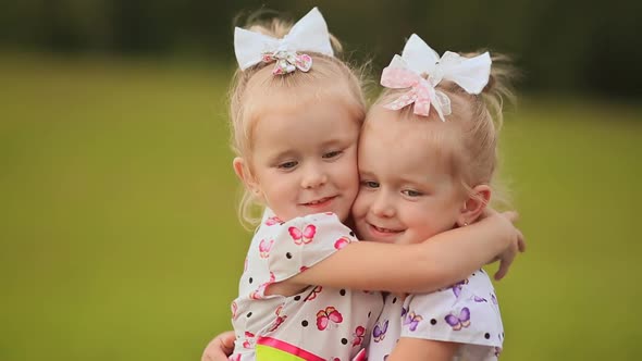 Two Little Twin Sisters are Pressed Against Each Other Having Fun on a Green Meadow in Summer