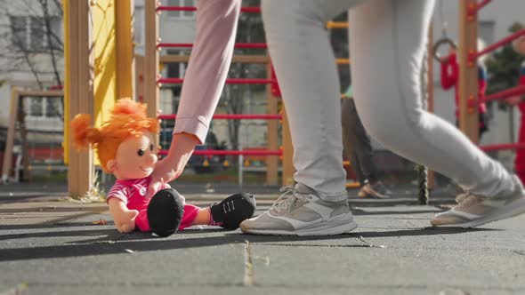 A toy doll sits on the ground on the playground. A woman passes by and takes a toy with her.
