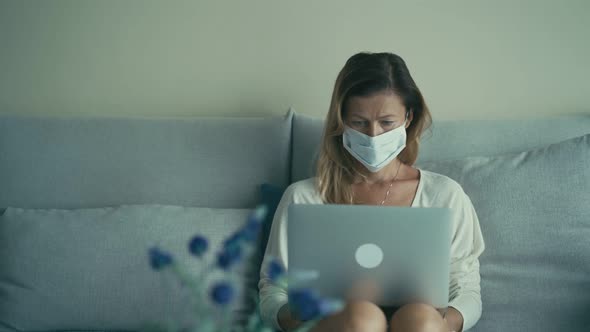 Business Woman in Disposable Medical Mask Is Typing on Laptop Keyboard on the Couch