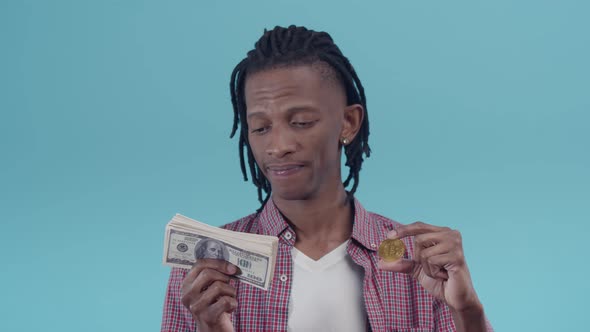 Black man with dreadlocks looks at a pack of dollar bills and a gold bitcoin coin in hands