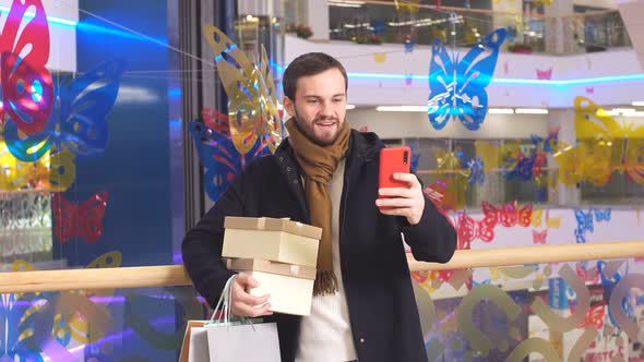 Happy Man with Gifts in Hands Talking on Skype with Interior of Shopping Center