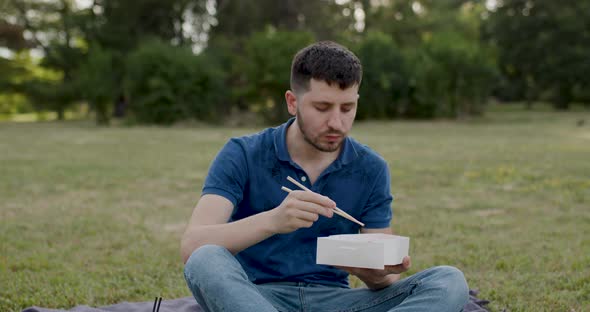 Man in Blue Tshirt Sitting on the Grass in the Woods Puts Sushi in His Mouth with Bamboo Sticks and