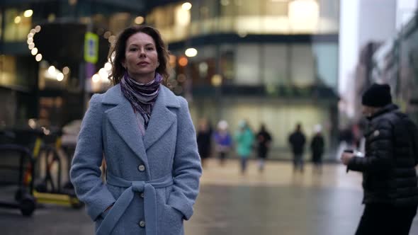 a Woman in a Light Coat and Scarf Walks Along a City Street at Dusk Against a Background of Blurred