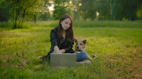 A Woman in a Park Next To an Open Laptop Is Petting Her Dog.