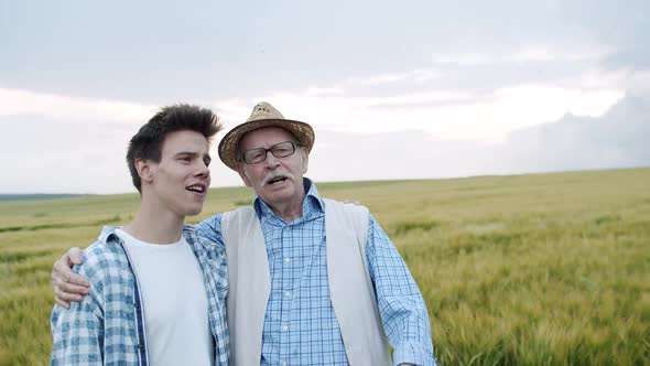 Happy Senior Farmer Stands in Embraces with Son in Barley Field and They Singing