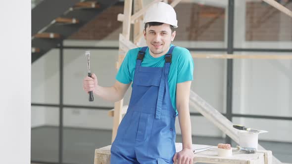 Positive Young Caucasian Builder Repairman Posing with Hammer in New Premises Under Construction