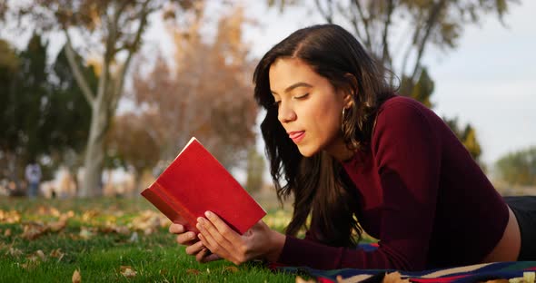 A hispanic woman college student reading a red book and smiling while laying down in the grass of a