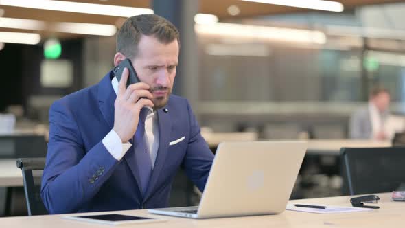 Middle Aged Businessman with Laptop Angry on Smartphone