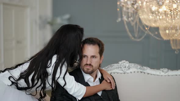 a Bride with Long Dark Hair is Hugging an Unshaven Groom in a White Shirt and Suit on a Light Sofa