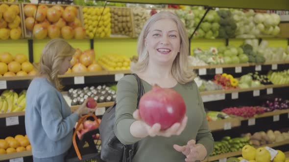 Cheerful Senior Caucasian Woman Stretching Pomegranate To Camera and Talking As Other Female