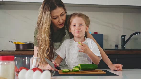Little Girl Eats Cucumber Slice Cooking Salad with Mother