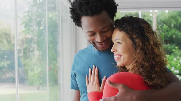 Happy biracial couple embracing, standing at window and looking into distance