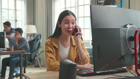 Asian Female Talking On Mobile Phone While Works On Her Personal Computer With Big Display In Office