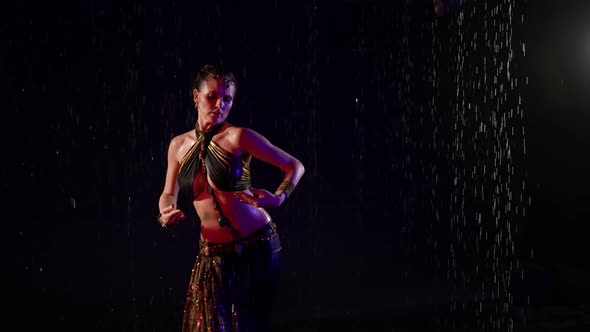 A Woman is in a Studio in the Pouring Rain on a Dark Background Dancing an Oriental Dance and Making