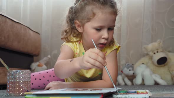 A little girl draws with watercolors on paper, lying on the floor at home. Art, education, childhood