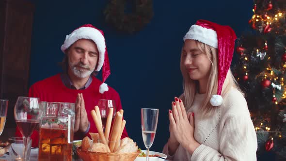 Young Woman and an Adult Man in Christmas Hats Sit at Table with Their Hands Folded and Pray Before