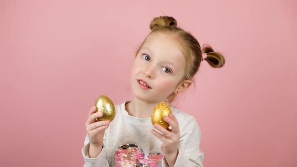 Funny Little Blonde Girl Smiling and Playing with Golden Easter Eggs on Pink Background