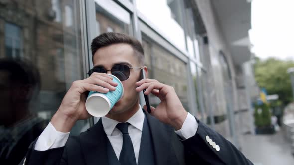 Happy Man in Sunglasses Greets with a Hand When Talks on Smartphone Outdoors