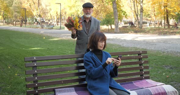 Elderly Woman Sits on a Bench and Uses a Smartphone, and Suddenly Her Husband Approaches Her, Gives