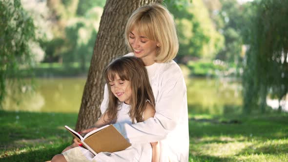 Happy Smiling Mother and Daughter Reading Book in Sunny Park Outdoors