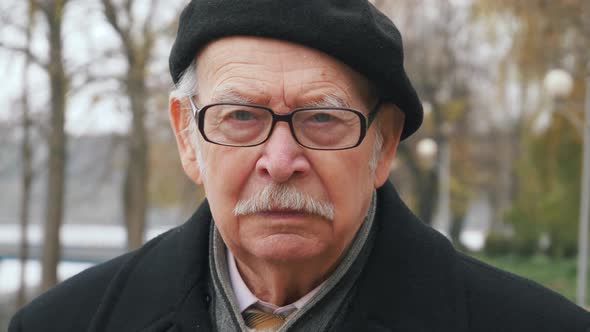 Portrait serious face caucasian old man looking at camera, outdoors close up.