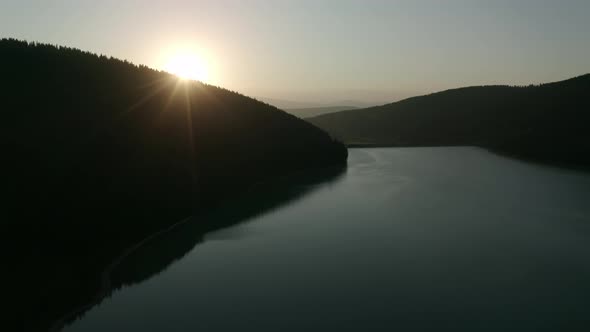 Slow aerial descent towards calm lake, sun setting behind ridge in Romania