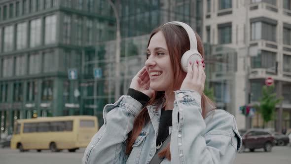 A Young Brunette Woman in Headphones is Listening to Music and Amusingly Dancing on a City