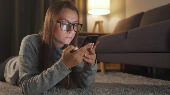 Woman with Glasses Is Lying on the Floor and Using Smartphone in the Evening