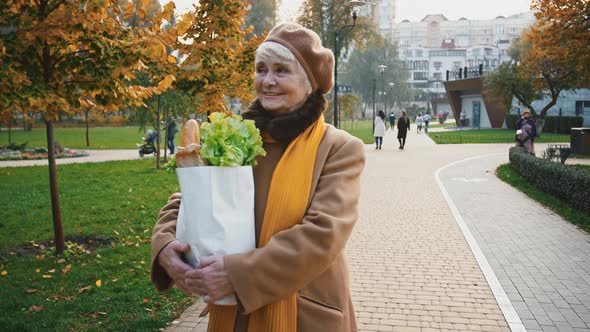 Aged Lady is Smiling and Holding Paper Bag of Groceries Baguette and Lettuce While Walking in Autumn