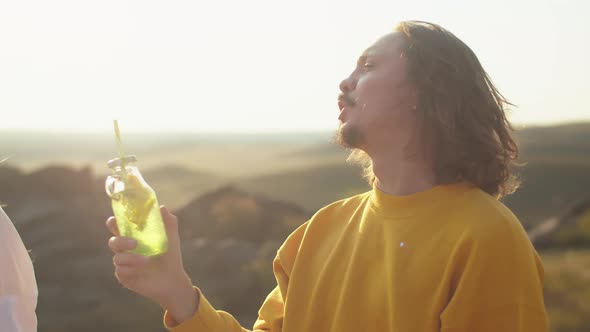 Man Drinks Cocktail Through a Straw and Talks to Woman in Nature Against the Background the Sun