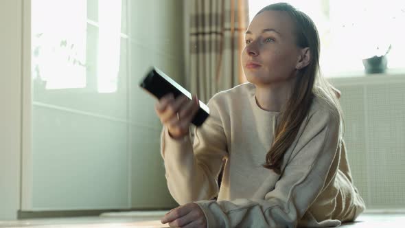 Young Smiling Woman with Tv Remote Control Watching the Television Lying on Living Room Floor in